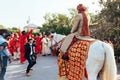 Indian groom ridding white horse with yellow and red pattern fabric, flower necklace and red turban with guests.