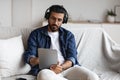 Indian guy spending time at home with digital tablet and wireless headphones Royalty Free Stock Photo