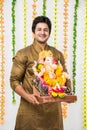 Indian handsome man in ethnic wear holding a Ganesh idol, welcoming God on Ganesh Chaturthi / festival at home with happy expressi