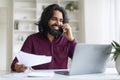 Indian man discussing work on phone while reviewing documents and using laptop Royalty Free Stock Photo