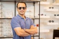 Indian man wear sunglasses with action of arm-crossed of confident in front of shelves with many types of glasses in optical shop