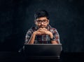 Indian programmer sits at a desk with a laptop and look at the camera with a thoughtful look.