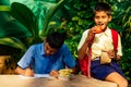 Indian school boy writing on notepad doing homework , looking concentration . His friend with backpack sits on table and Royalty Free Stock Photo