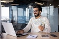 Indian smiling young man businessman working in the office on a laptop, sitting at the table and holding cash bills in Royalty Free Stock Photo