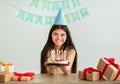 Indian teenage girl in hat holding birthday cake with candles, smiling at camera, sitting at table with gifts at home Royalty Free Stock Photo