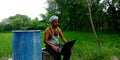 An Indian village farmer operating laptop computer system seating at water pot in agriculture wheat grain grass field area