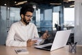 Indian young male businessman working in office, sitting at desk holding notebook and talking on video call via laptop Royalty Free Stock Photo