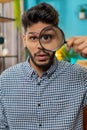 Indian young man holds magnifying glass near face looking at camera with big zoomed eye analyzing Royalty Free Stock Photo