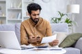 Indian young man working from home, sitting on sofa and checking bills and documents, crumpling papers in hands, looking Royalty Free Stock Photo