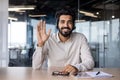 Indian young smiling businessman sitting in the office at the desk in front of the camera in the headset, waving and Royalty Free Stock Photo