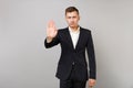 Indifferent young business man in classic black suit, shirt showing stop gesture with palm isolated on grey wall
