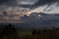 Indonesian landscape with rainy sky on sunset - dark blue clouds with orange, yellow sunbeams over hazy mountains, smoky green.