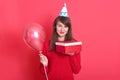 Indoor picture of magnetic lovely young woman having holiday cap on head, holding red baloon and red and white present box, Royalty Free Stock Photo