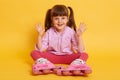 Indoor picture of playful energetic active little girl raising hands, sitting on floor isolated over yellow background, wearing