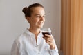 Indoor portrait of a beautiful young dark haired woman standing with a glass of red wine and smiling, posing in light living room Royalty Free Stock Photo