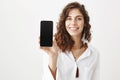 Indoor portrait of charming young woman with curly hair smiling broadly while holding smartphone, advertising gadget