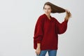 Indoor portrait of curious romantic caucasian woman in loose red sweater, holding beautiful healthy hair in hand aside