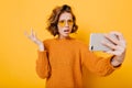 Indoor portrait of disappointed short-haired girl in glasses making selfie in studio. Upset young woman in knitted