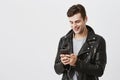 Indoor portrait of handsome caucasian guy in trende leather jacket holding mobile phone, messaging with his friends Royalty Free Stock Photo