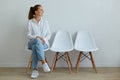 Indoor portrait of young attractive woman sitting on chair in waiting room, wearing jeans and white t shirt, looking away with Royalty Free Stock Photo