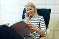 Indoor portrait of young beautiful blonde woman talking on mobile phone, sitting on comfortable chair and reading magazine in the Royalty Free Stock Photo