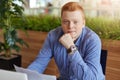 Indoor portrait of young stylish businessman with red hair dressed in checked shirt sitting a office working trough papers, studyi Royalty Free Stock Photo