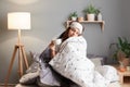 Indoor shoot of sleepy woman wearing blindfold sitting on bed wrapped in blanket, holding cup of coffee, having nap, posing with