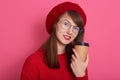 Indoor short of attractive woman in red beret, sweater anfd eyewear, holding cup of coffee or tea in hands isolated over pink Royalty Free Stock Photo