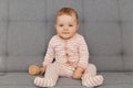 Indoor shot of adorable cute toddler baby girl wearing striped sleeper playing with her wooden toy alone while sitting on sofa, Royalty Free Stock Photo