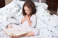 Indoor shot of adorable girl reading book while sitting in bed, cute child looks in book with great interest and keeps hands on