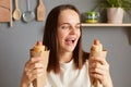 Indoor shot of amazed excited hungry woman with brown hair wearing white t-shirt, holding two hot dogs in hands, keeps mouth open