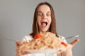 Indoor shot of amazed excited woman with brown hair taking big plate with tasty pasta isolated over gray background, being