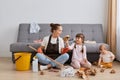 Indoor shot of angry Caucasian woman wearing white t shirt, brown apron and jeans, cleaning house, posing with her children near