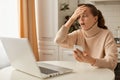 Indoor shot of astonished scared woman wearing beige sweater posing in kitchen, looking at cell phone screen with open mouth, Royalty Free Stock Photo