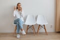 Indoor shot of attractive astonished woman wearing white shirt and jeans sitting on chair in queue, talking phone and raising