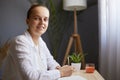 Indoor shot of attractive beautiful smiling young woman writing down notes while sitting at table in office, looking at camera Royalty Free Stock Photo