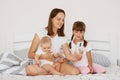 Indoor shot of attractive Caucasian female wearing white casual style t shirt sitting on bed with her two daughters, holding her Royalty Free Stock Photo