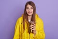 Indoor shot of attractive lady standing against lilac studio wall, holding thermo mug with hot beverage, girl wearing yellow Royalty Free Stock Photo