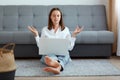 Indoor shot of attractive woman wearing white shirt and jeans sitting on floor and working on laptop, being tired, trying to relax