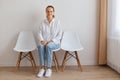 Indoor shot of attractive young adult woman sitting on chair against white wall, wearing shirt and jeans, posing with smile, Royalty Free Stock Photo