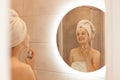 Indoor shot of beautiful girl in bath towel on her head using perfume while posing in bathroom, enjoying pleasant odor, looking at Royalty Free Stock Photo