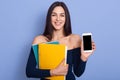 Indoor shot of beautiful winsome woman posing isolated over blue background, holding two paper folders and mobile phone with blank Royalty Free Stock Photo