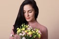 Indoor shot of beautiful young girl with long brunette hair, dressed shirt, holding spring flowers, looking at her bouquet with