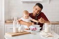 Indoor shot of brunette man in maroon t shirt and apron sitting at table with infant daughter in kitchen, talking phone with Royalty Free Stock Photo