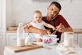 Indoor shot of busy father sitting at table with baby daughter in hands and cooking in kitchen, talking phone with sad expression Royalty Free Stock Photo