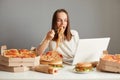 Indoor shot of busy hungry woman eating fast food while working on laptop, sitting at table against gray wall, working on laptop