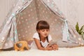 Indoor shot of calm pretty little girl with braids wearing white t shirt lying in wigwam with smart phone in hands, watching Royalty Free Stock Photo