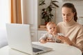 Indoor shot of Caucasian dark haired woman wearing beige sweater sitting at table with baby girl in hands and using cell phone, Royalty Free Stock Photo