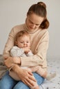 Indoor shot of Caucasian dark haired woman with ponytail wearing beige sweater sitting on bed and hugging her little toddler Royalty Free Stock Photo