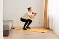 Indoor shot of Caucasian women wearing white t shirt and black leggins doing sports exercise, doing squats with dumbbells in hands Royalty Free Stock Photo